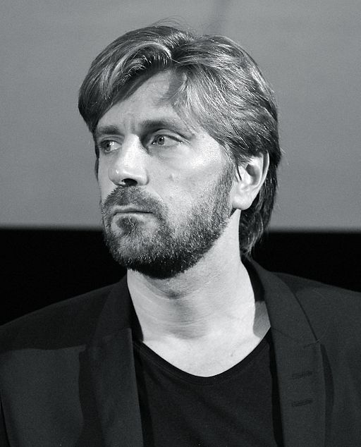 Ruben Östlund sns från axlarna ch uppåt. Han har halvlångt hår, skägg ch svart kavaj.