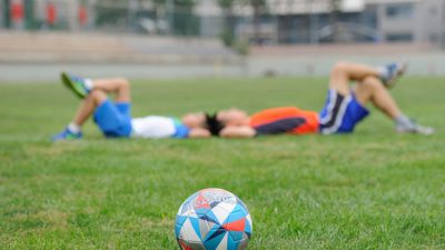 En fotboll ligger på en gräsmatta. I bakgrunden ligger två barn med huvudena mot varandra och benen korsade.