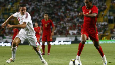 En fotbollsspelar i rött springer med bollen. Bredvid honom springer en spelar i vitt och försöker jaga ikapp honom. Bakom dem syns en stor fotbollsplan och en stor läktare.