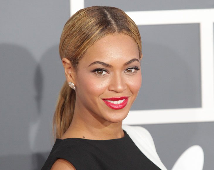 Beyonce står framför en grå vägg. Hon har blonderat hår i en mittbena och hästsvans. Hon har klarrött läppstift och svarta kläder.