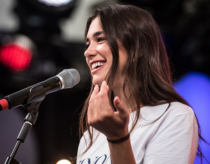 Dua Lipa står på en scen framför en mikrofon. Hon har långt brunt hår och en vit t-shirt. Hon ler stort.