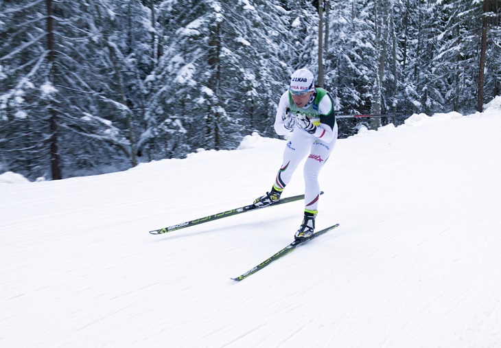 Charlotte åker skidor fort fram genom en snötäckt skog. Hon har färgglada skidor och vita tajta kläder. Hon har mössa, gula glasögon och håller stavarna under armarna så de pekar bakåt.