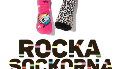 Två fötter sticker in i bilden uppifrån. Ena foten har en rosa strumpa med en enhörning. Den andra foten har en leopardfärgad strumpa. Under fötterna står det med stora bokstäver "Rocka sockorna".