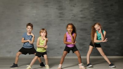Fyra barn står uppställda i shorts och kortärmat. De står med benen brett isär, böjda knän och håller ett finger på kinden. De ser ut att vara mitt i en dans, eller precis har avslutat en.