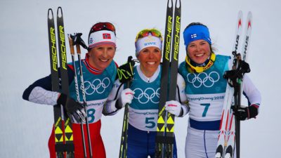 Björgen, Kalla och en till skidåkare står bredvid varandra. De har tajta skidkläder och nummerlappar på magen. De håller i var sitt par med längdskidor. Alla ler stort och tittar in i kameran.