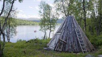 En kåta står i skogen intill en sjö. Kåtan ser ut som ett spetsigt tält, men gjort av trä.