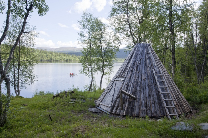 En kåta står i skogen intill en sjö. Kåtan ser ut som ett spetsigt tält, men gjort av trä.