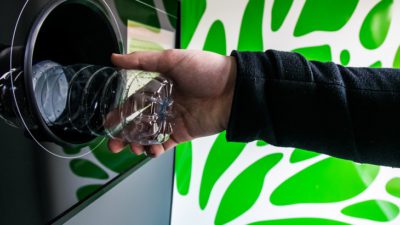 En arm sträcker fram en plastflaska mot en pantmaskin. Flaskan försvinner halvvägs in i ett hål i maskinen.