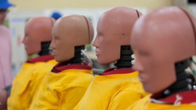 Flera stora dockor sitter i rad. De har huvuden med ansikten som liknar människor, men utan ögon. Nacken är en svart fjäder. De har gula rockar på sig.