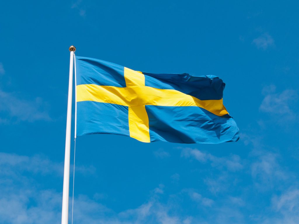En svensk flagga vajar i vinden på en flaggstång. Solen lyser på flaggan och himlen är blå bakom.