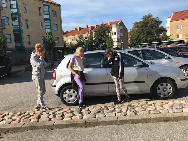 Tre barn håller för munnen vid en bil på en parkering.