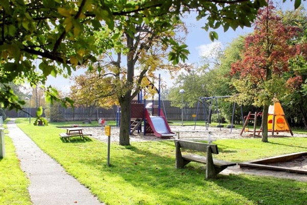 Lekplats med gungor och rutchkana, träd, bänkar och sandlåda.
