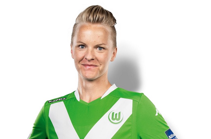 Nilla står mot en vit vägg och ler mot kameran. Hon har en grön fotbollströja och har kort hår som är bakåtkammat.