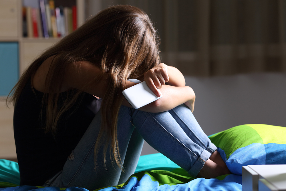 En tonåring sitter på en säng. Personen sitter med uppdragna knän, huvudet vilar mot knäna och den kramar om sina ben. I handen håller personen en telefon.